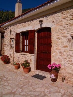 A stone building with a red door and window shutters, potted plants, and a metal grate on the ground. - Arhontiko Kordopati Traditional Guesthouse (Tripoli)