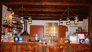 A rustic bar area with wooden chairs, a counter with various bottles and a menu, and a large window in the background. - Arhontiko Kordopati Traditional Guesthouse (Tripoli)
