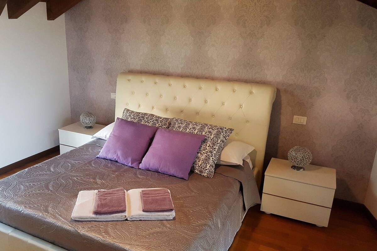 A bedroom with a tufted headboard, patterned bedding, two bedside tables with decorative items, and a window with blinds.