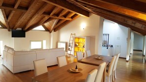 A spacious loft with a high ceiling, wooden beams, a dining area with a wooden table and chairs, a white sofa, and a ladder leading to a lofted storage area. - Donizetti Royal (Bergamo)