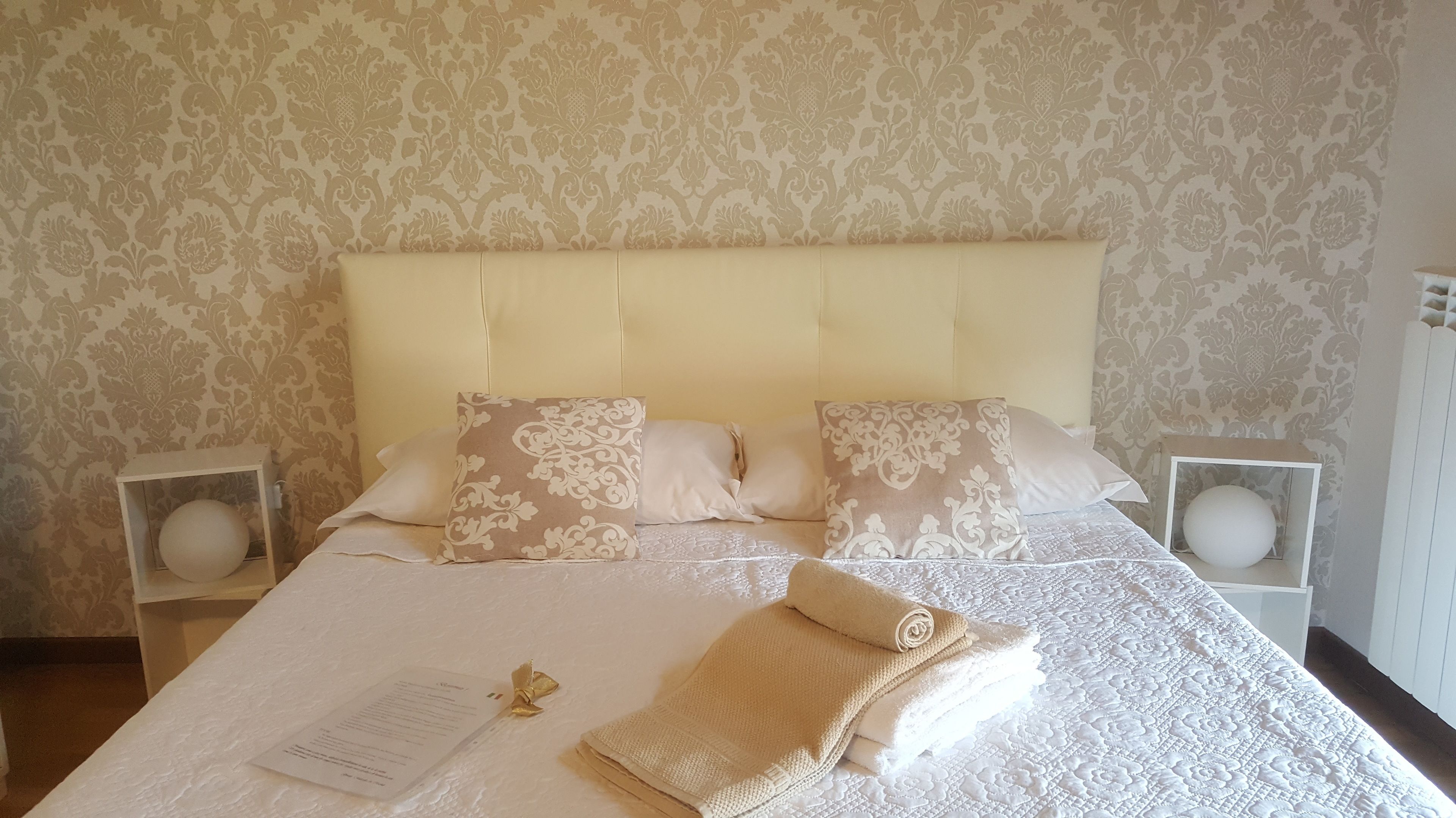 A neatly made bed with a white and beige patterned bedspread, two decorative pillows, a folded towel, and a small white table with a round object on it.