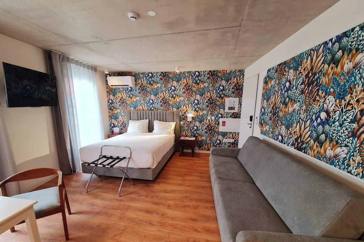 A hotel room with a bed, a grey sofa, a wooden chair, a small table, and a wall with a floral pattern.