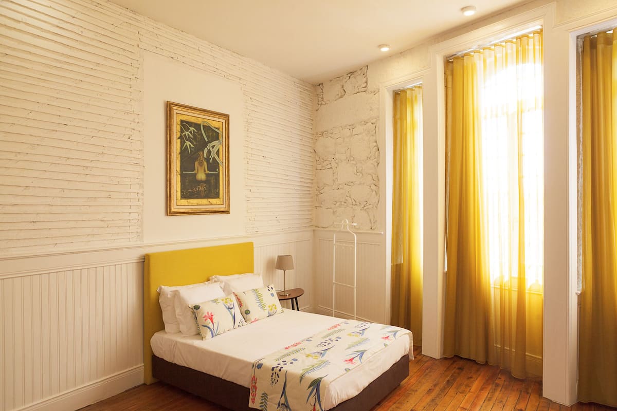 A bedroom with a bed, a nightstand, a lamp, a window with curtains, and a painting on the wall.