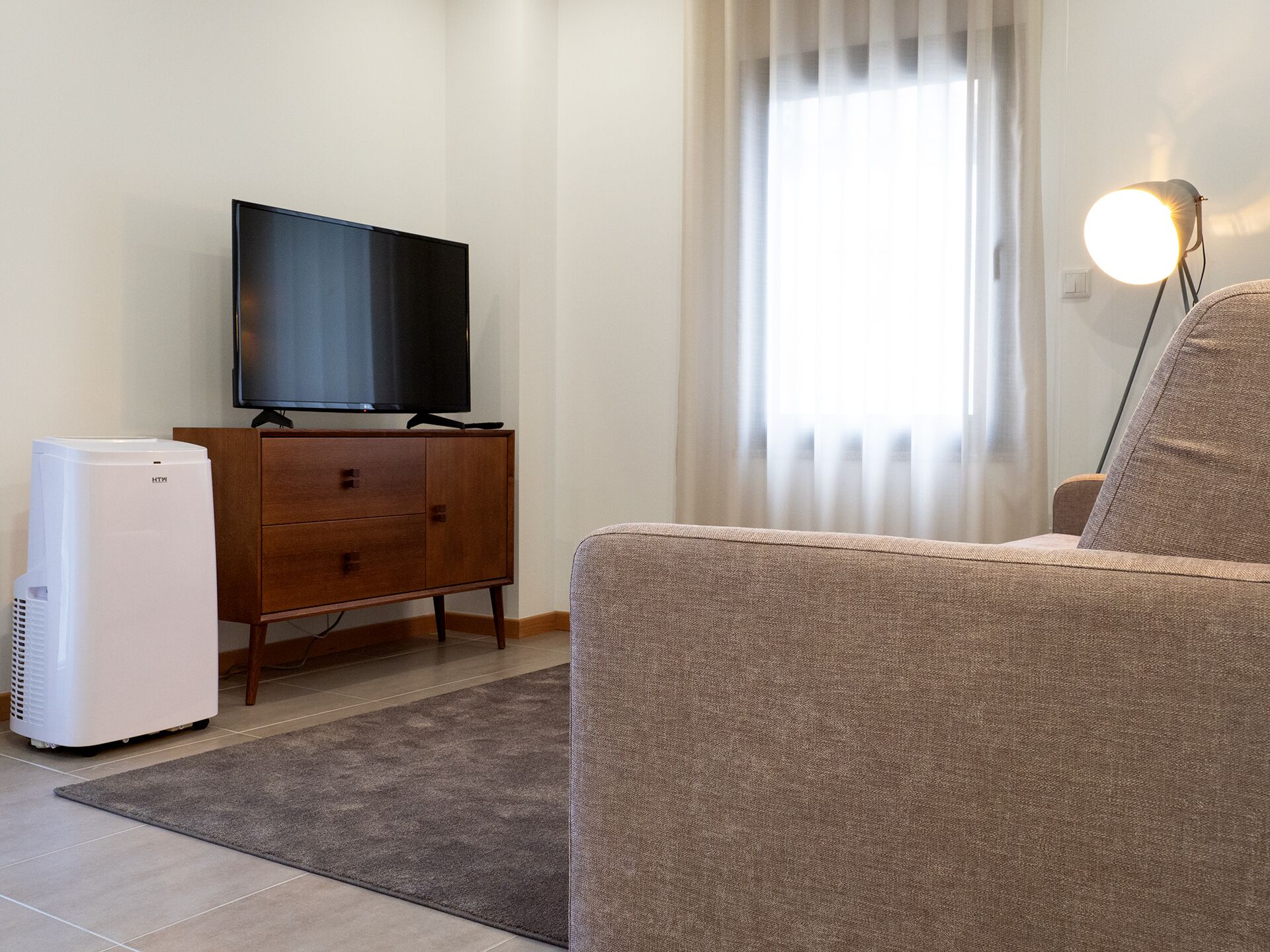 A living room with a flat-screen TV on a wooden cabinet, a white air conditioner, a brown sofa, and a lamp with a round shade.