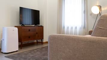 Una sala de estar con un televisor de pantalla plana sobre un mueble de madera, un aire acondicionado blanco, un sofá marrón y una lámpara con pantalla redonda.