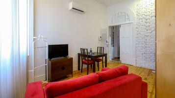 Un espacio habitable compacto con un sofá rojo, un televisor sobre un mueble de madera, una mesa de comedor con sillas y una pared de ladrillo blanco.