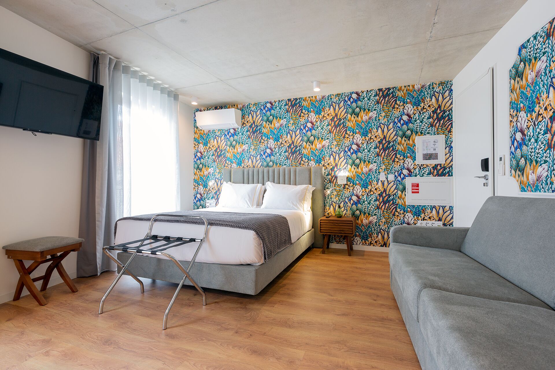 A modern hotel room with a bed, a grey sofa, and a wooden floor.