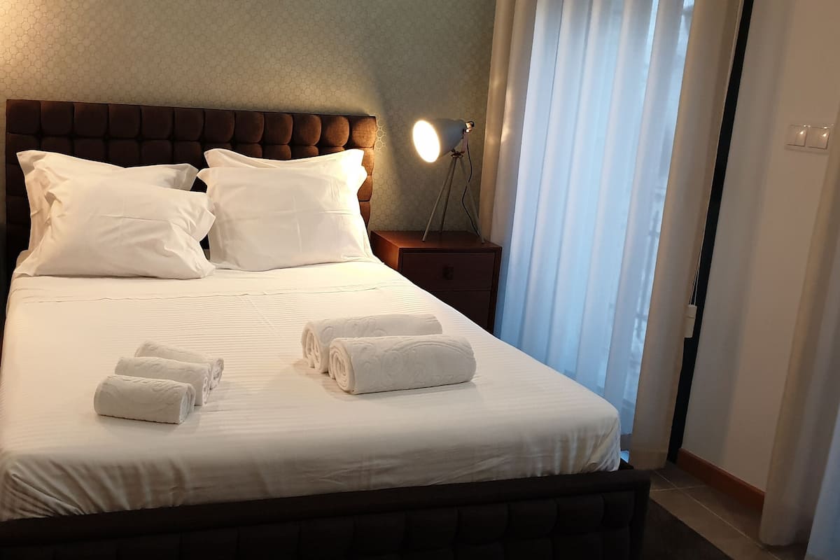 A neatly made bed with white linens and pillows, a bedside lamp, and rolled towels on the bed.