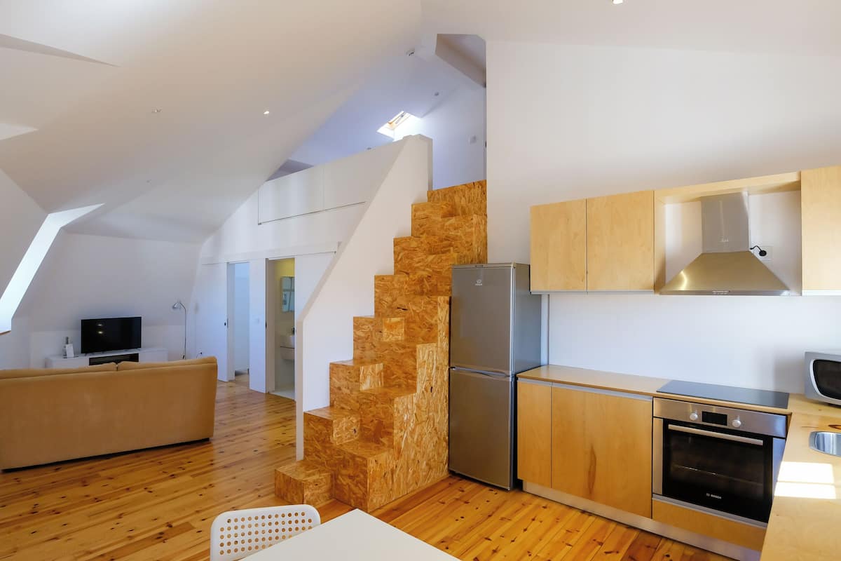 A modern kitchen with wooden cabinetry, a built-in oven, and a refrigerator. There is a wooden staircase leading to an upper level.