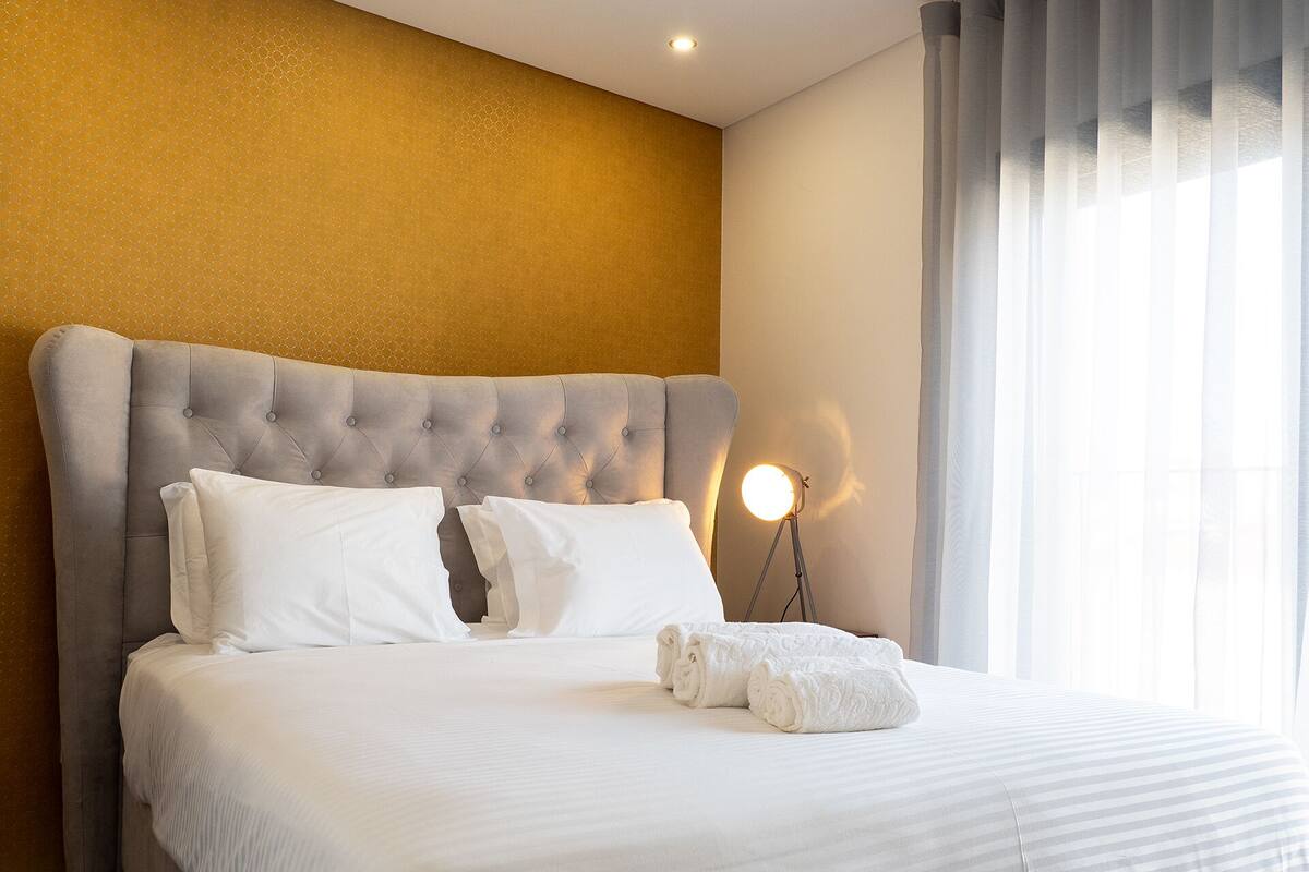 A neatly made bed with white linens and pillows, a tufted headboard, and a bedside lamp.