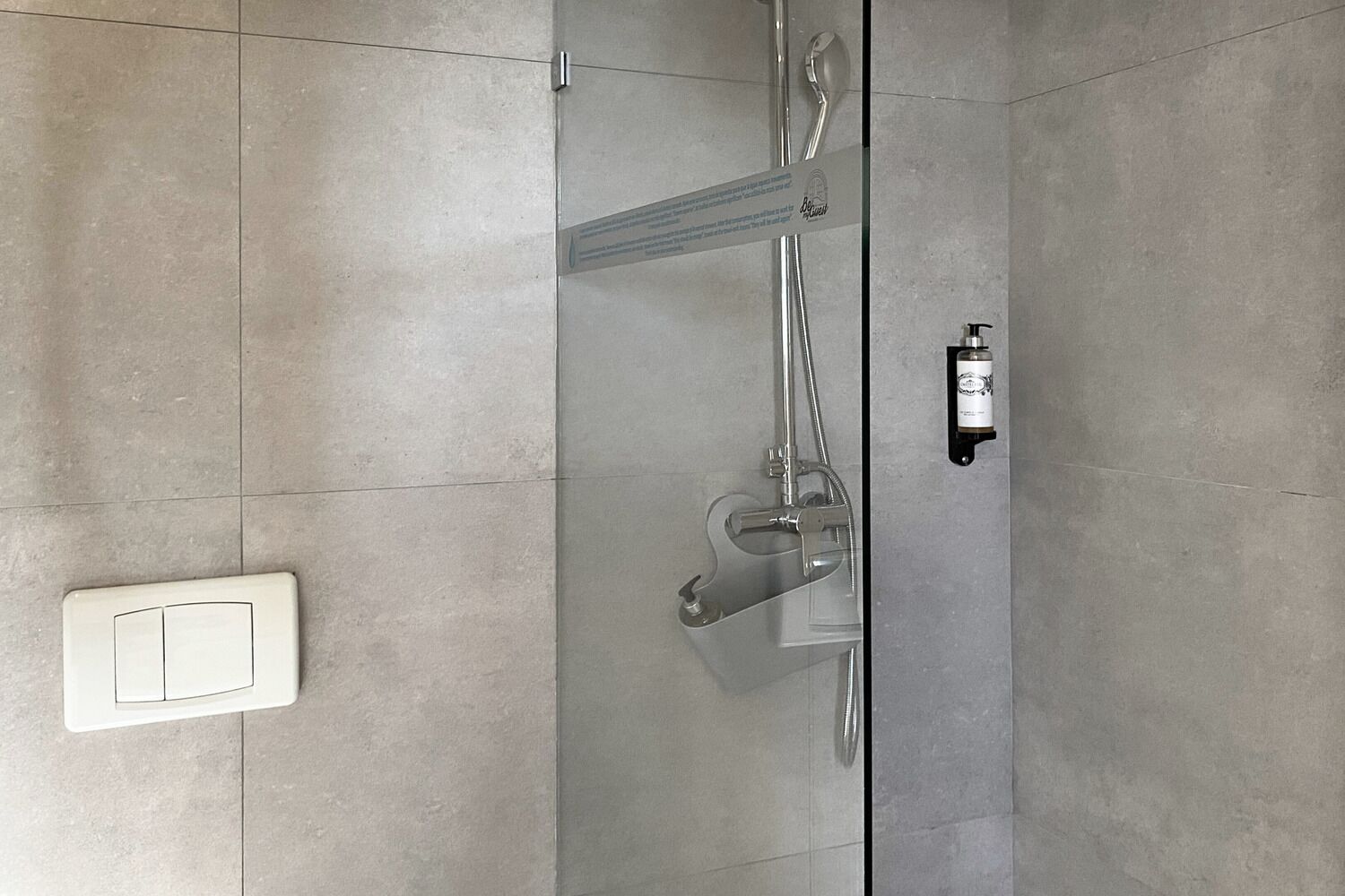 Una ducha moderna con puerta de vidrio, cabezal de ducha de mano y una botella de champú en la pared.