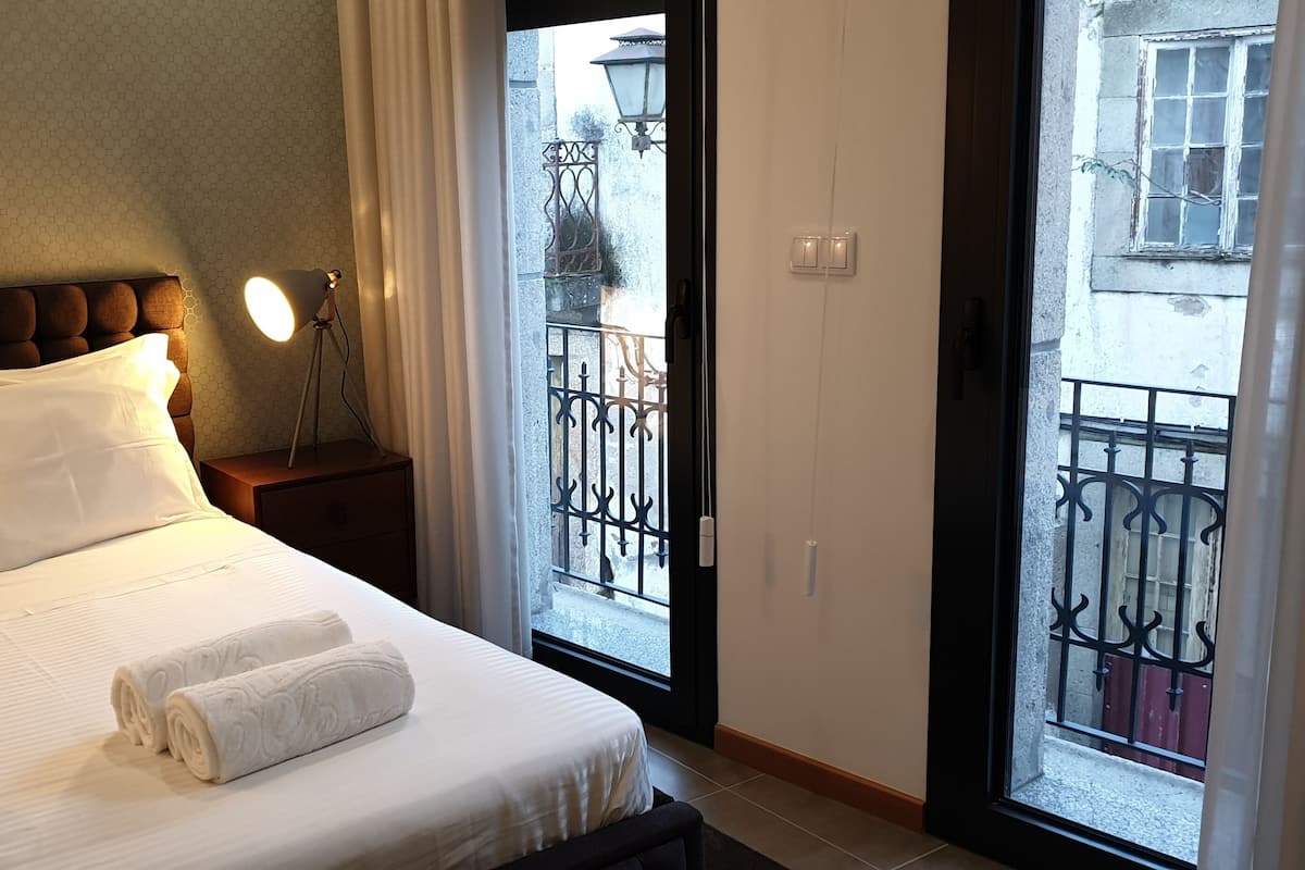 A hotel room with a bed, white linens, a nightstand with a lamp, and a view of a balcony with a wrought iron railing.