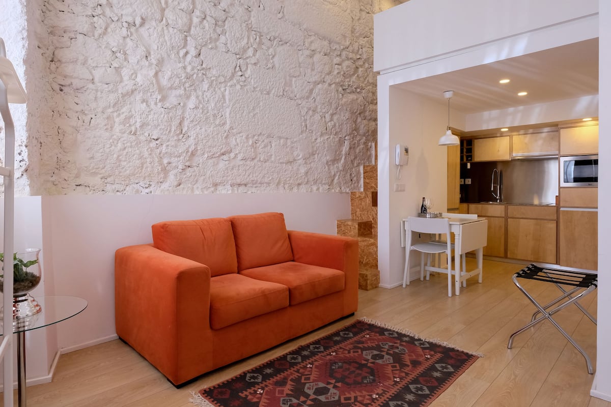 A modern living room with a sofa, a glass table with a plant, a patterned rug, and a kitchen area with wooden cabinets and a stainless steel refrigerator.