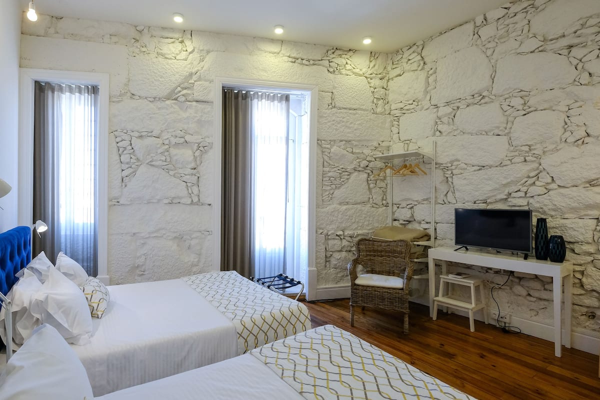 A hotel room with two beds, a desk with a TV, a wicker chair, and stone wall decor.