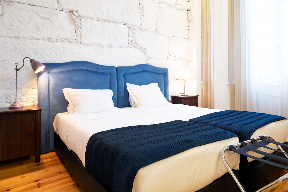A bed with a blue headboard, white bedding, and a dark blue blanket. There is a nightstand with a lamp and a small table with a lamp. The room has a stone wall and a window with sheer curtains.