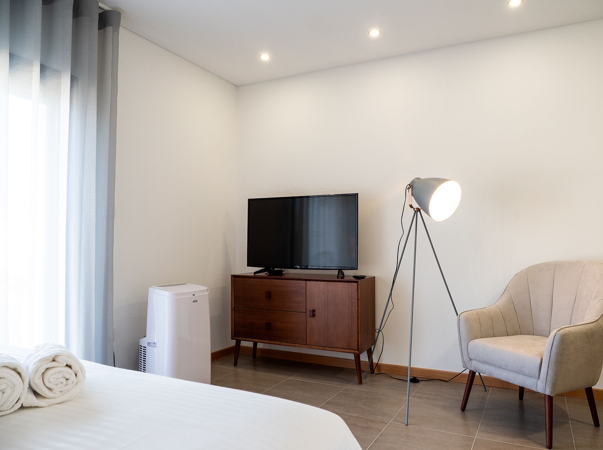 Habitació d'hotel amb un llit, un televisor sobre un moble de fusta, una làmpada de peu i un aire condicionat blanc.