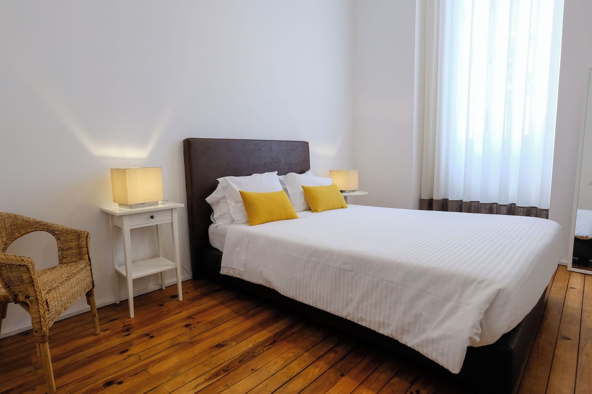 A bedroom with a wooden floor, a bed with white bedding and two yellow pillows, a wicker chair, and two bedside tables with lamps.