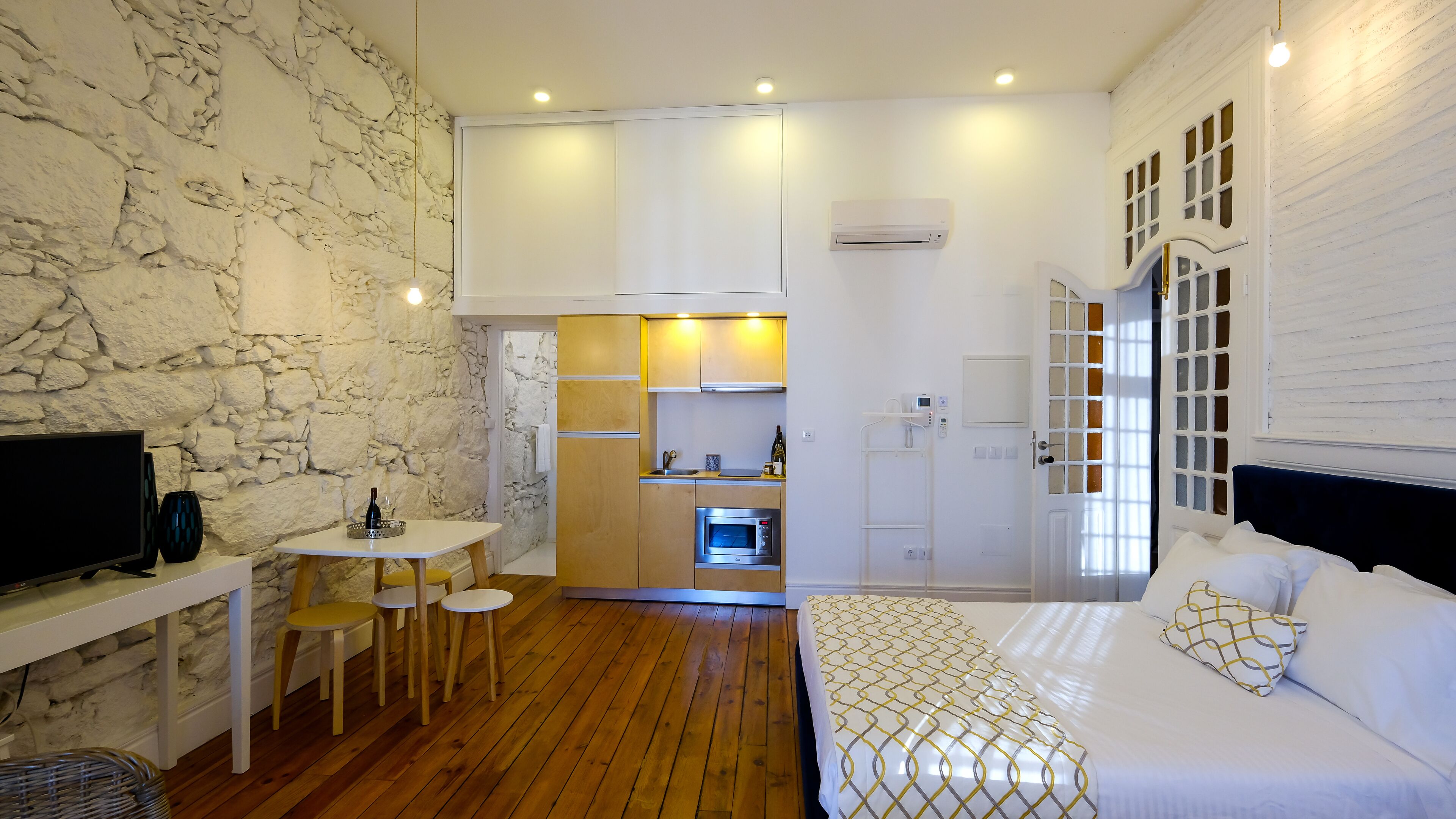 A compact hotel room with a stone wall, a wooden floor, a bed with patterned bedding, a small kitchenette, and a television on a desk.