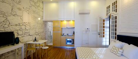 A compact hotel room with a stone wall, a wooden floor, a bed with patterned bedding, a small kitchenette, and a television on a desk.