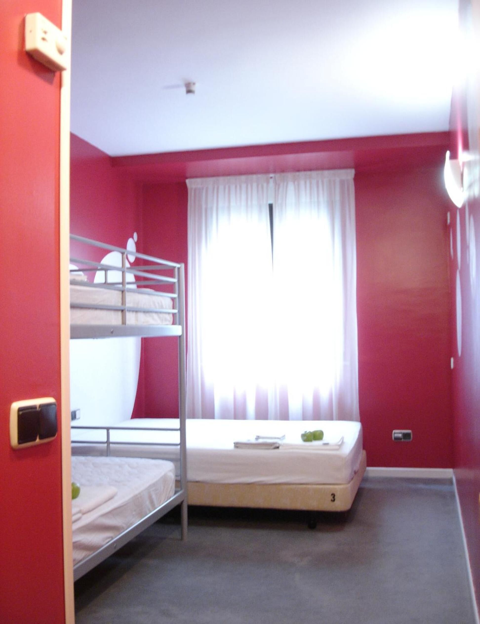 Une pièce aux murs rouges, avec un lit superposé, un lit simple, une fenêtre ornée de rideaux blancs et un interrupteur sur le mur.