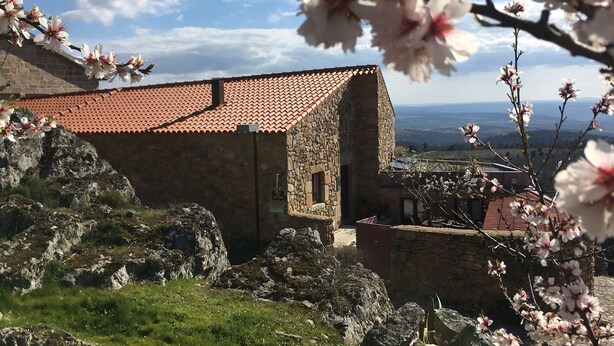 Un edificio de piedra con techo de tejas rojas, rodeado de rocas y vegetación, con vistas a colinas distantes.