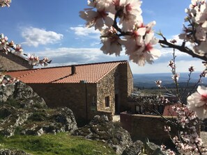 Un edificio de piedra con techo de tejas rojas, rodeado de rocas y vegetación, con vistas a colinas distantes.