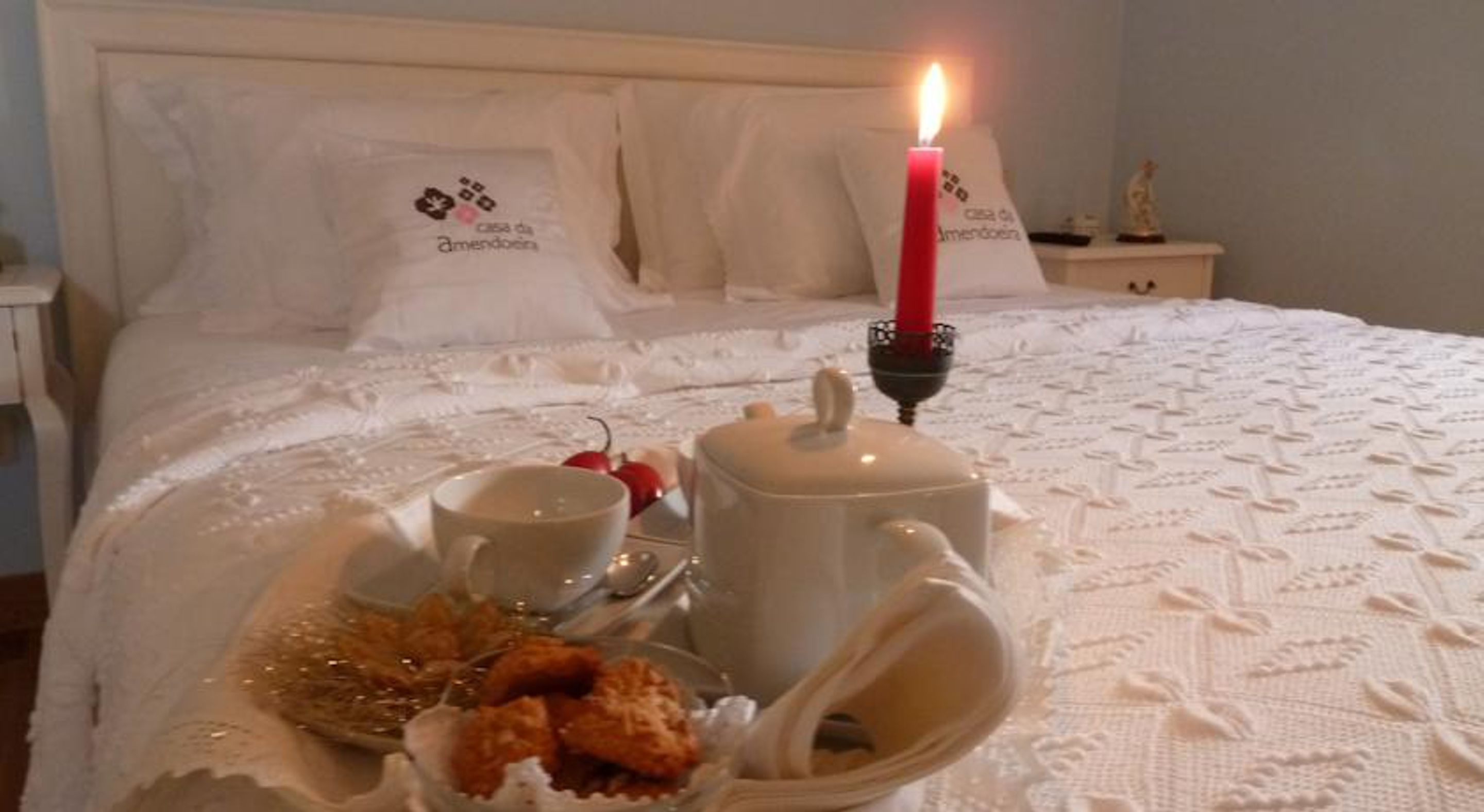 Una cama bien hecha con sábanas y almohadas blancas, una vela encendida, y una bandeja con tetera, tazas y comida.