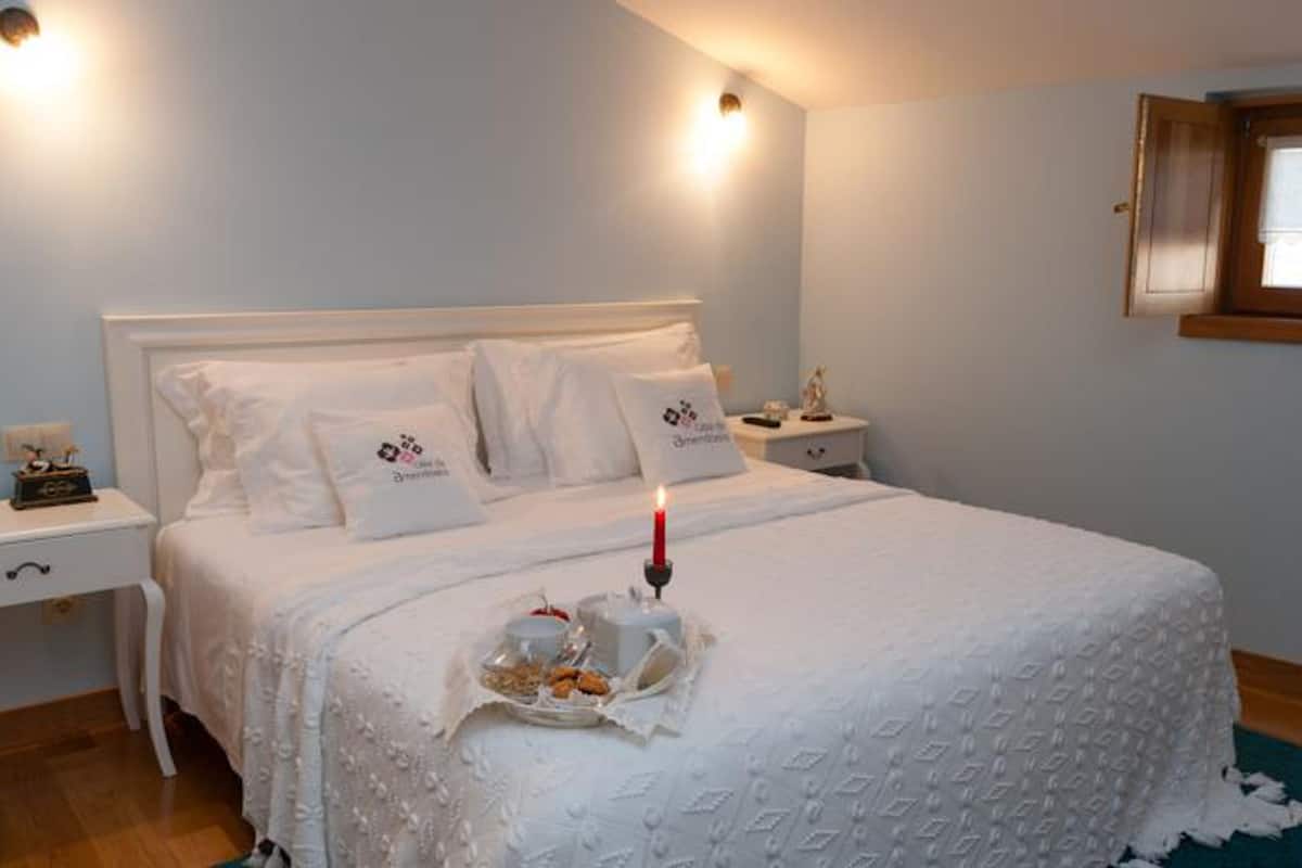 Una cama bien hecha con sábanas y almohadas blancas, una mesita de noche con una lámpara y una ventana con marco de madera.