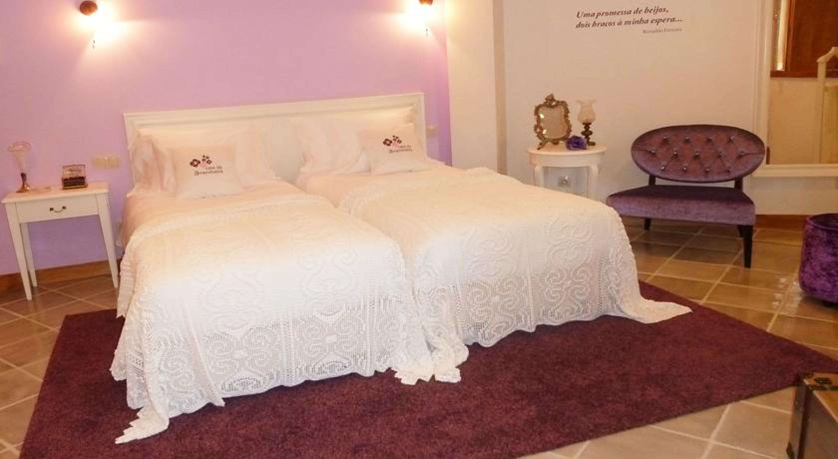 Un lit double avec du linge de lit blanc et une tête de lit assortie, une petite table de chevet avec une lampe, et un fauteuil violet dans une pièce dont le sol est carrelé et où se trouve un tapis violet.