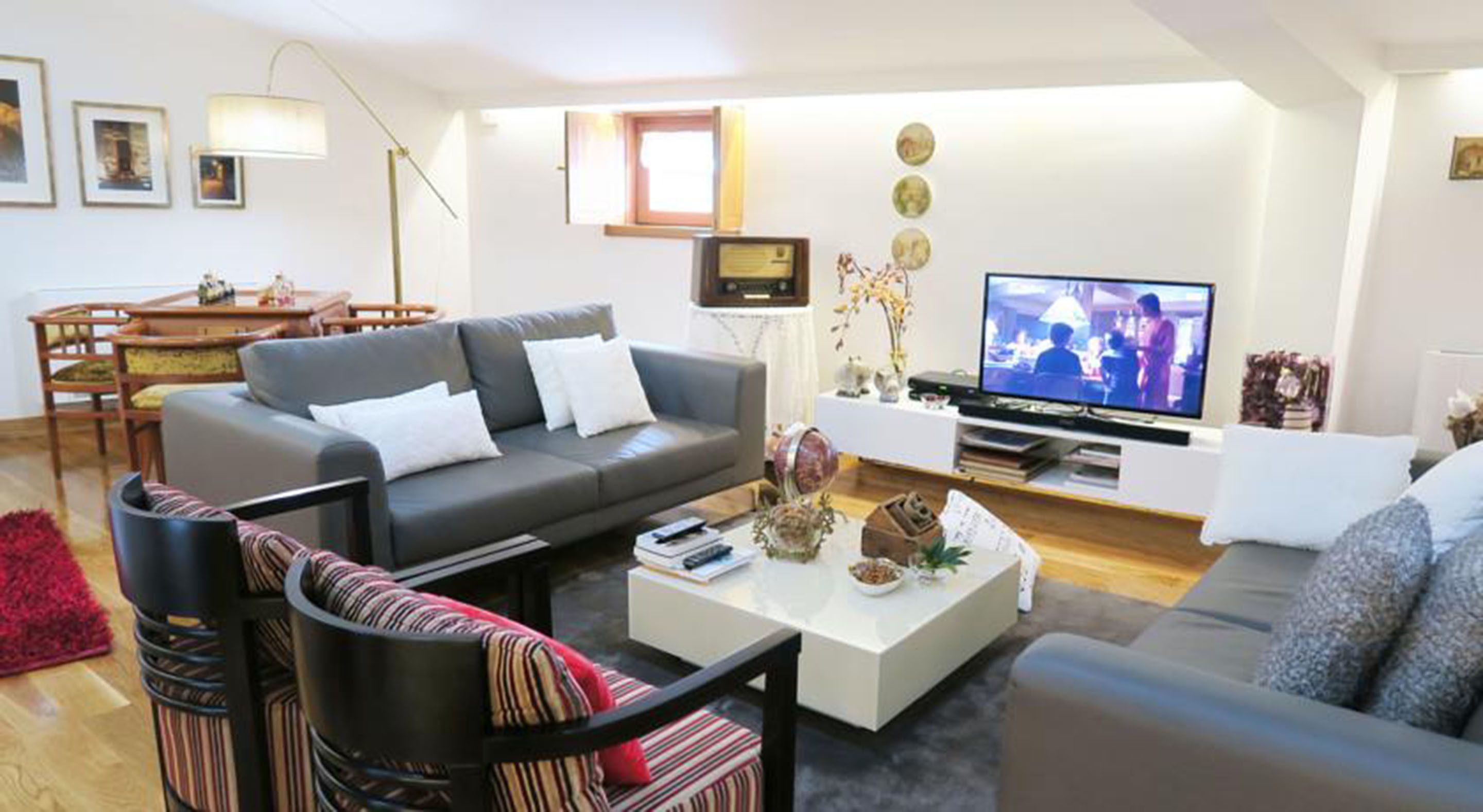 Una sala de estar moderna con un sofá gris, una mesa de centro con objetos decorativos, un televisor y un comedor con mesa y sillas de madera.