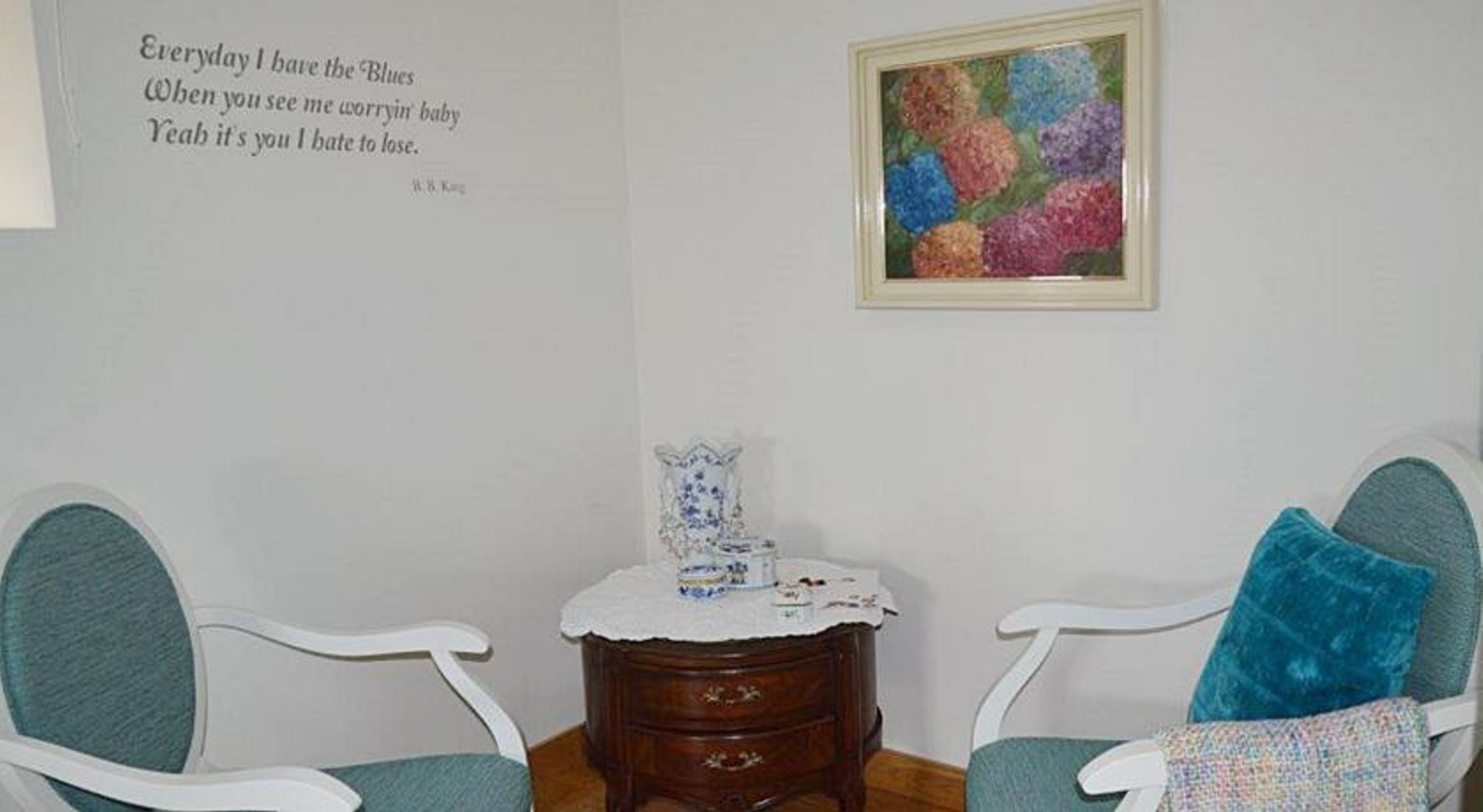 A room with two white chairs, a small wooden table with a decorative item, and a colorful painting on the wall.