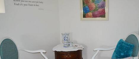 A room with two white chairs, a small wooden table with a decorative item, and a colorful painting on the wall.