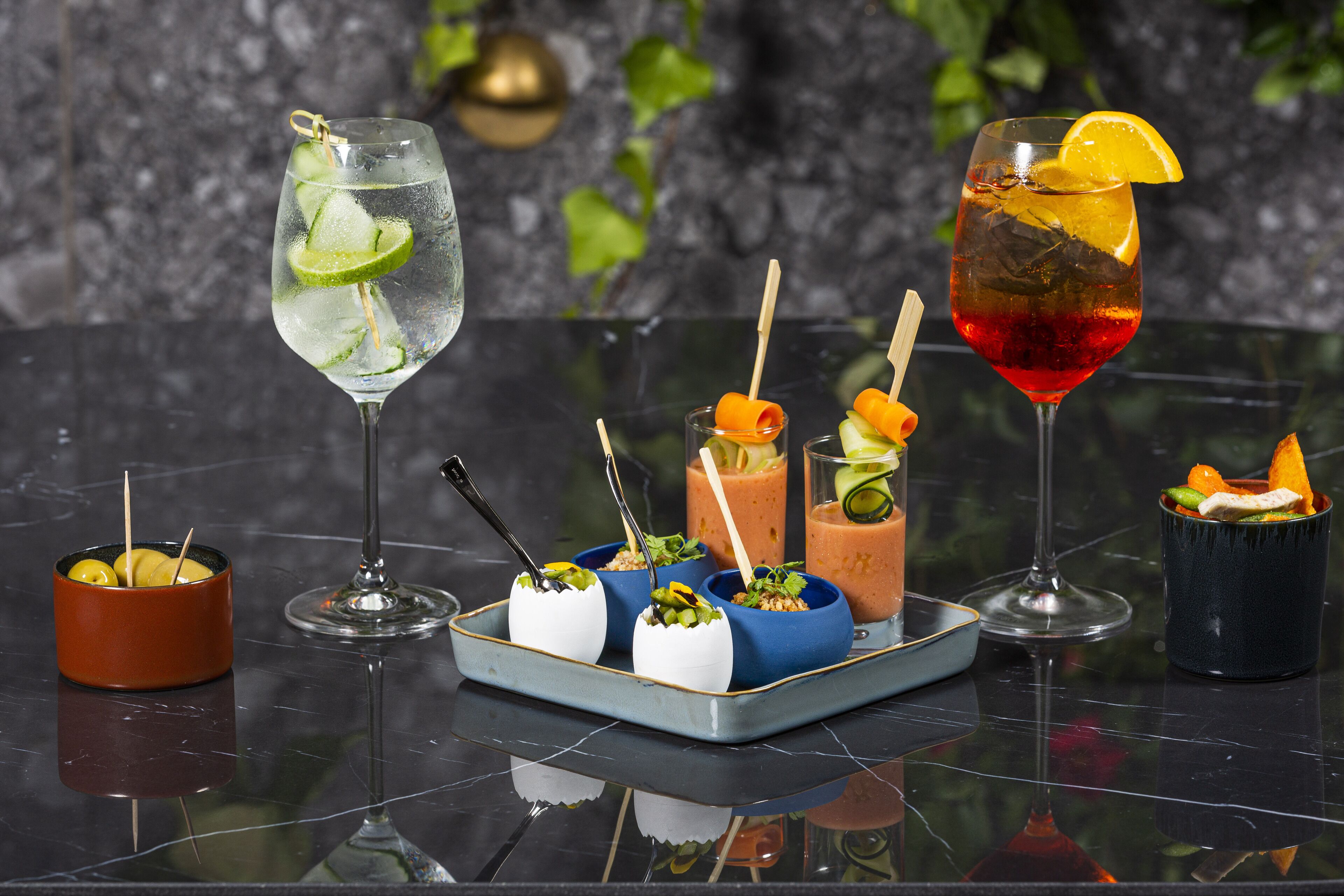 A variety of drinks and appetizers on a reflective surface, with a dark, textured background and green foliage.