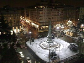 Μια νυχτερινή θέα μιας πλατείας πόλης, με ένα κεντρικό μνημείο, τα γύρω κτίρια και έναν πολυσύχναστο δρόμο με οχήματα.