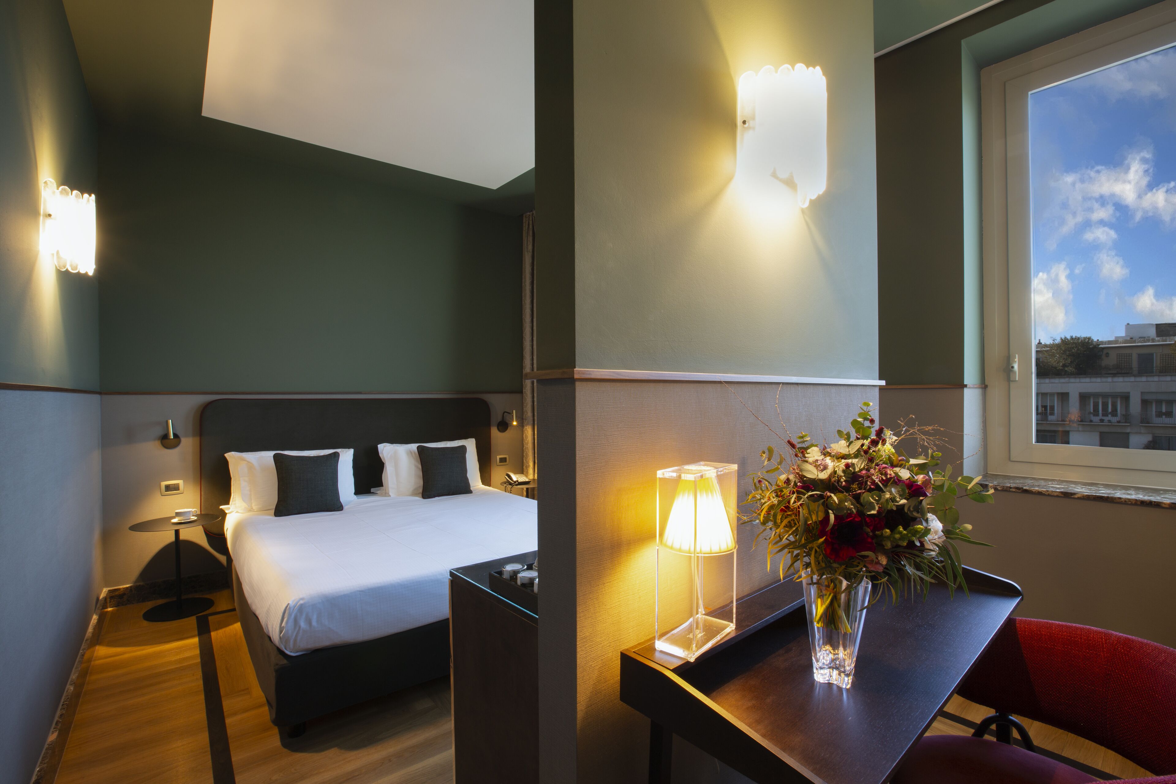 A hotel room with a bed, bedside table, lamp, and a vase with flowers.