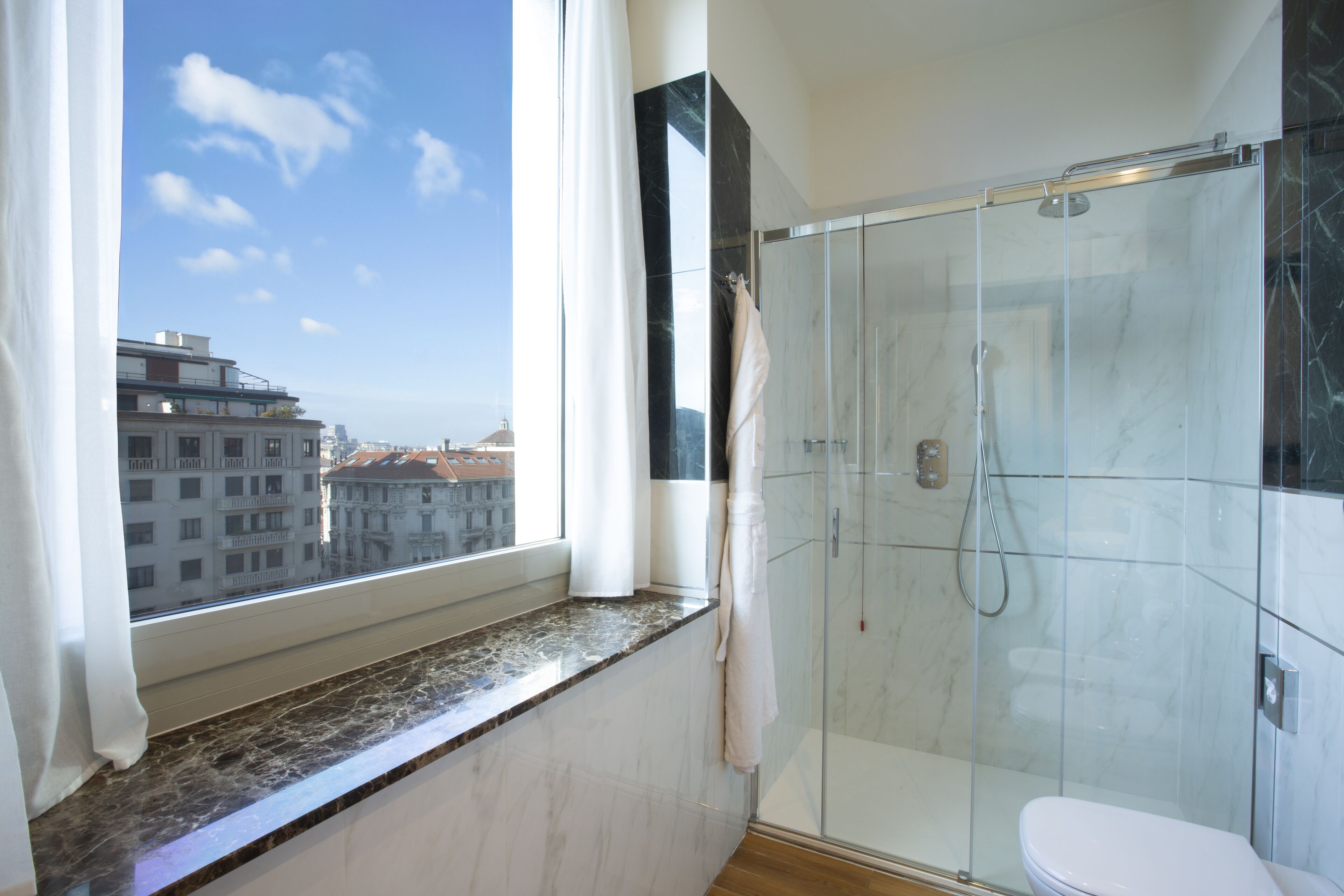 Un baño moderno con ducha de vidrio, inodoro y vistas a la ciudad a través de un ventanal amplio.
