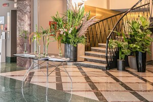 A lobby with a checkered marble floor, a glass table with magazines, and a staircase with potted plants.