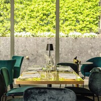 A dining area with a table set for two, surrounded by chairs, with a view of greenery through large windows.