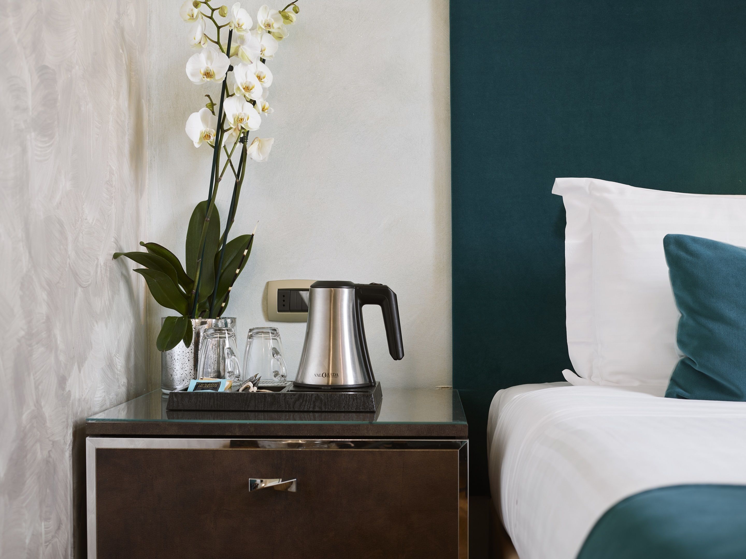Habitación de hotel con una cama, una mesita de noche con cafetera, un florero con flores blancas y un reloj de pared.