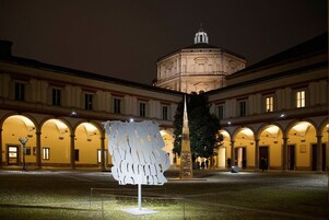 A courtyard with a modern sculpture, a historic building with arches, and a tall monument.