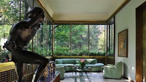Un interior moderno con una escultura grande, un sofá verde y vistas a árboles a través de amplios ventanales.