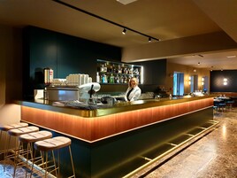 A modern bar area with a bar counter, stools, a bartender, and a well-stocked shelf with bottles.