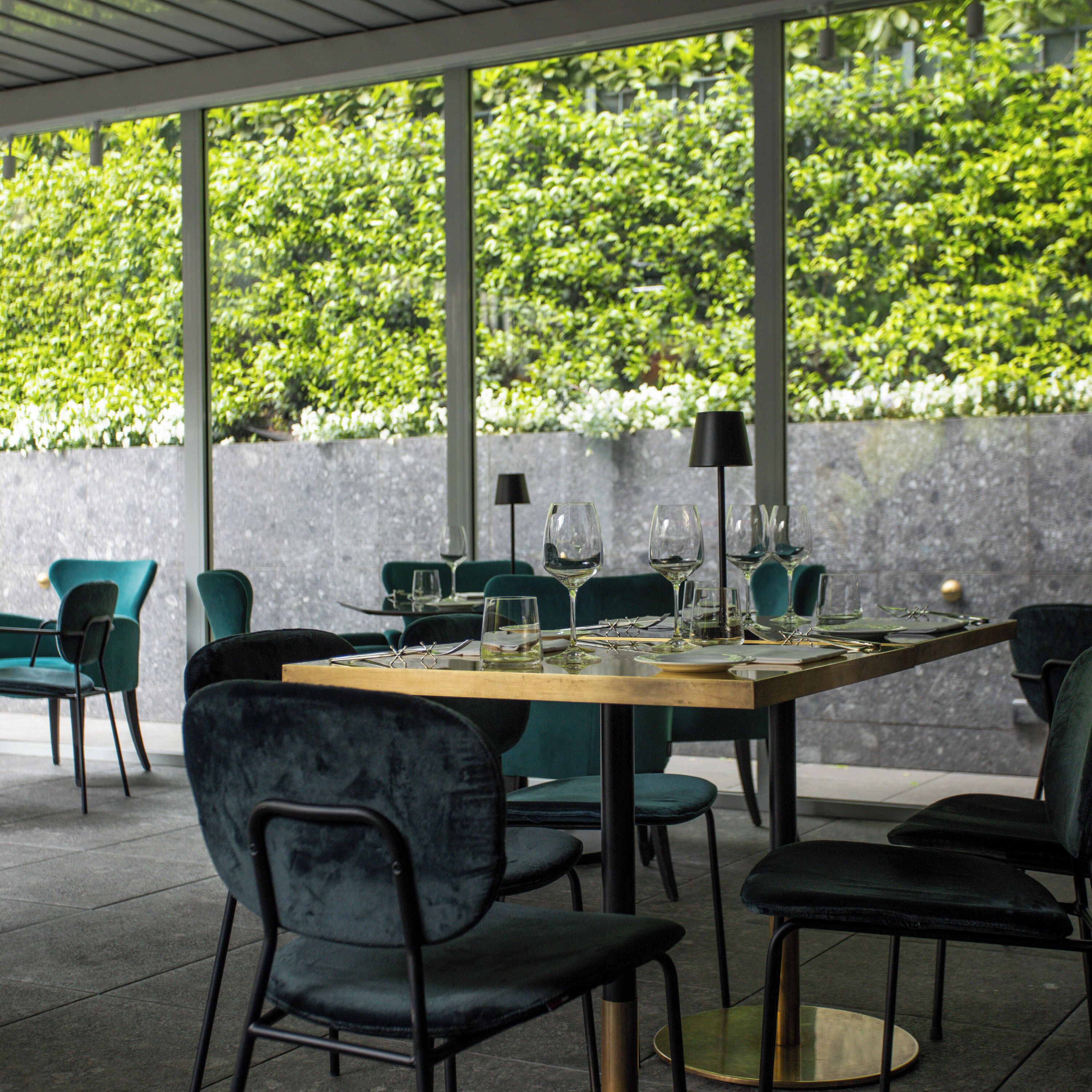 An outdoor dining area with a table set for two, surrounded by chairs, and a view of a lush green hedge.