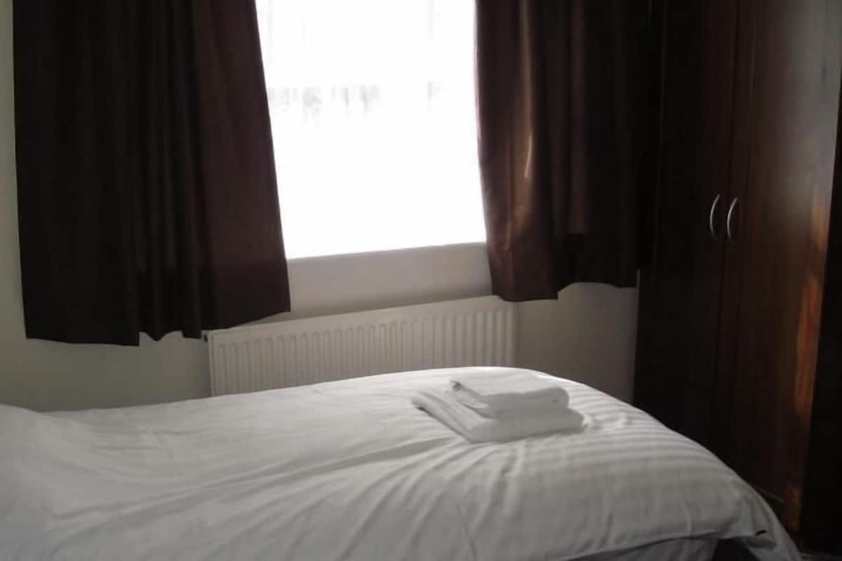 A hotel room with a bed, white bedding, a radiator, and dark curtains.