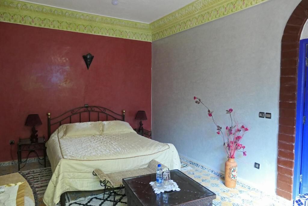 A hotel room with a bed, a small table with a bottle, a vase with flowers, and a patterned floor.