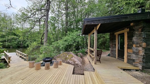 A wooden deck with a fire pit, a stone building with a black roof, and a wooden bridge over a river. - Villa Struka (Pyhtaa)