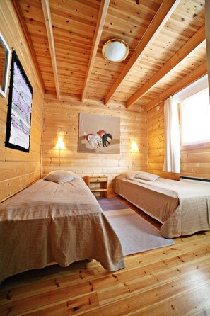 A cabin with two single beds, wooden walls, a ceiling with beams, a mounted painting, and a window with curtains.