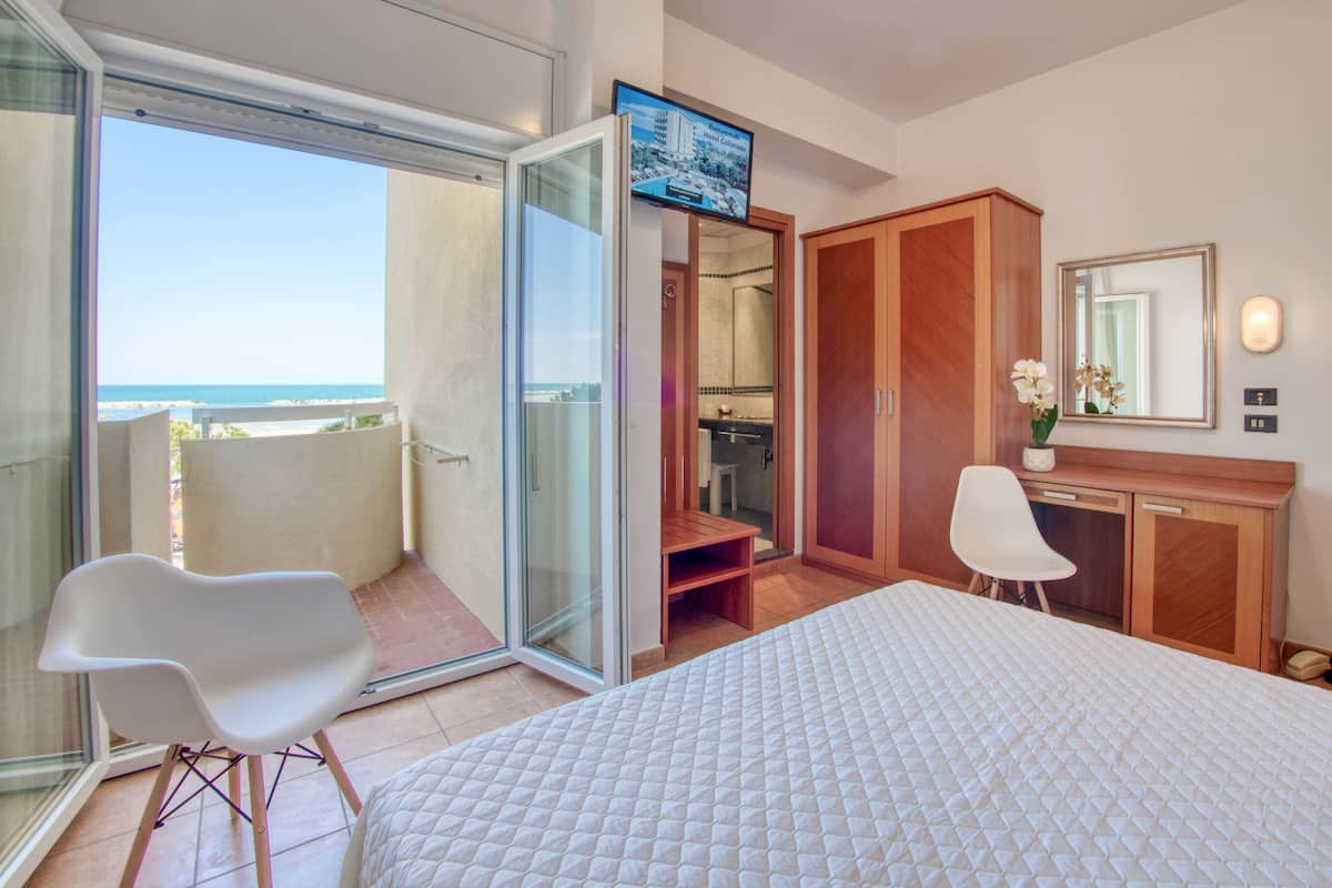 A hotel room with a bed, a desk, a chair, a wardrobe, and a sliding glass door leading to a balcony with a view of the ocean.