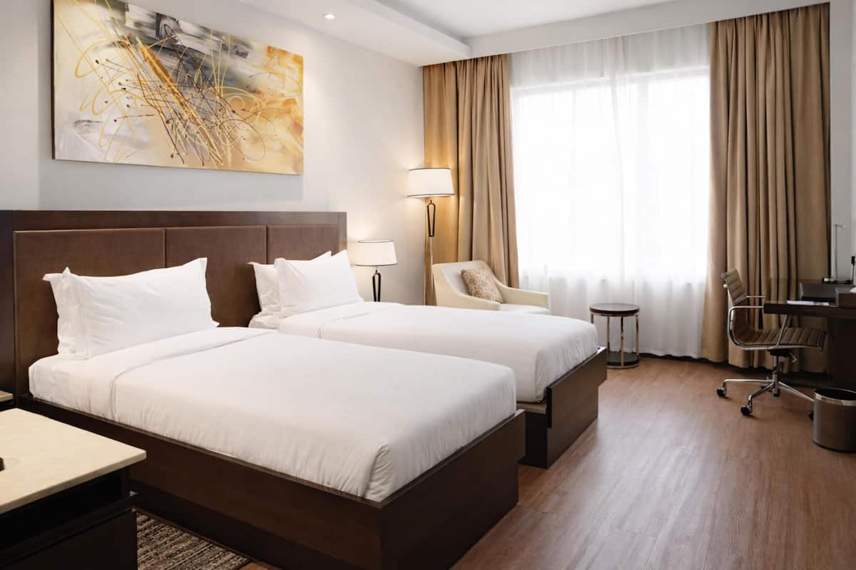 Ein Hotelzimmer mit zwei Betten, einem Schreibtisch, einem Stuhl, einer Lampe und einem großen Fenster mit Vorhängen.