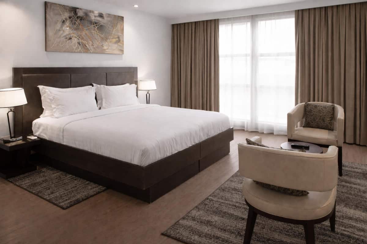 Ein Hotelzimmer mit einem großen Bett, zwei Nachttischen, einem Sessel, einem Schreibtisch, einer Lampe und einem Bild an der Wand.